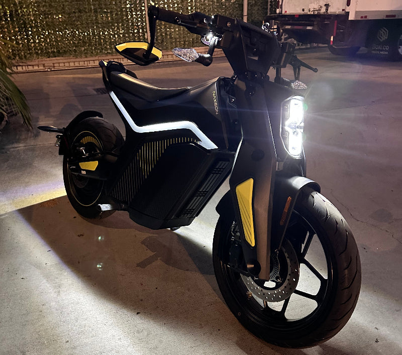 Black and yellow Naxeon electric motorcycle with LED headlight, parked on concrete at dusk