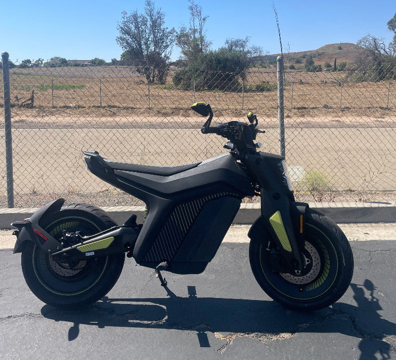 Black electric motorcycle with green accents parked on asphalt near chain-link fence and dry landscape