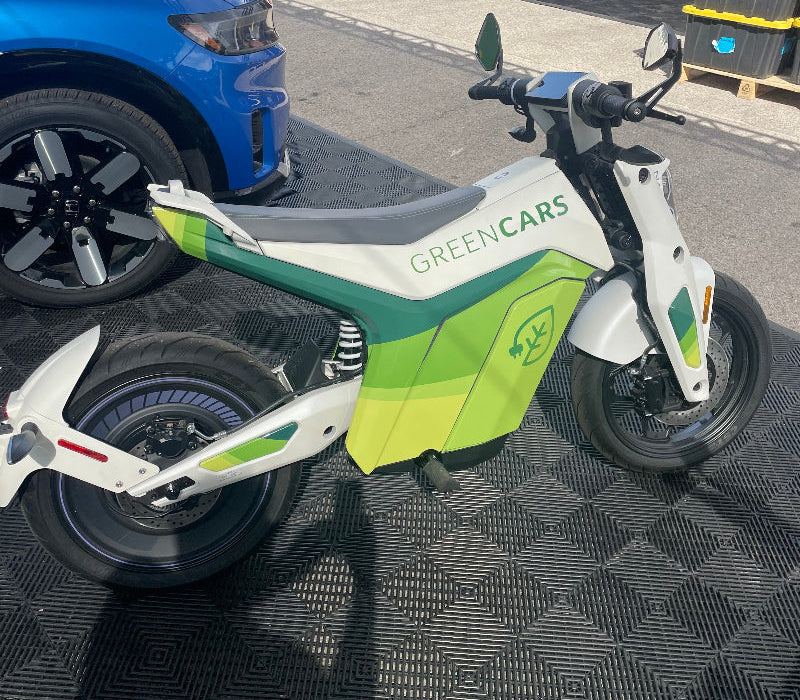Green and white electric motorcycle with GREENCARS logo parked on textured black floor at outdoor event