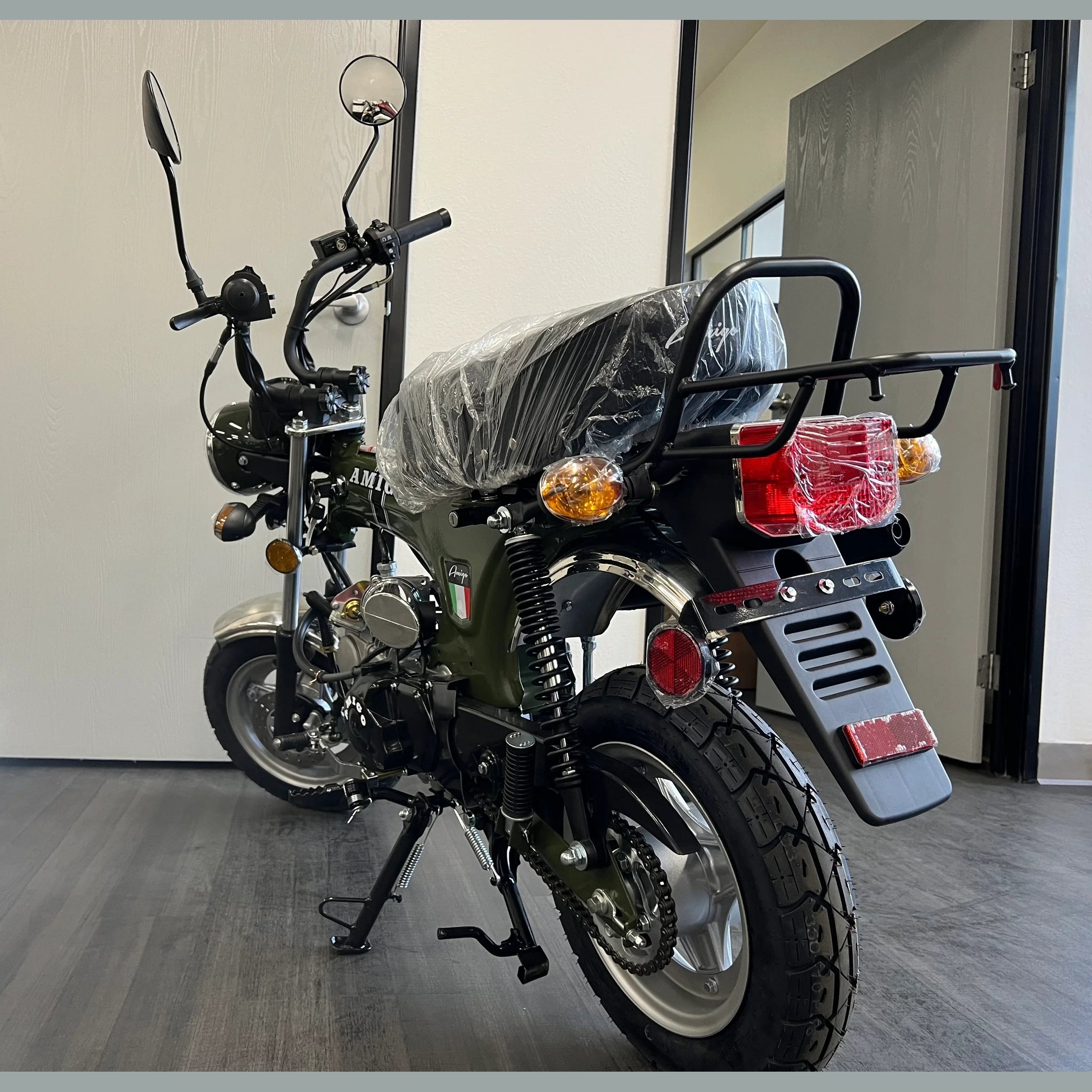 Green Amigo 125cc mini motorcycle rear view with knobby tires and plastic-wrapped seat