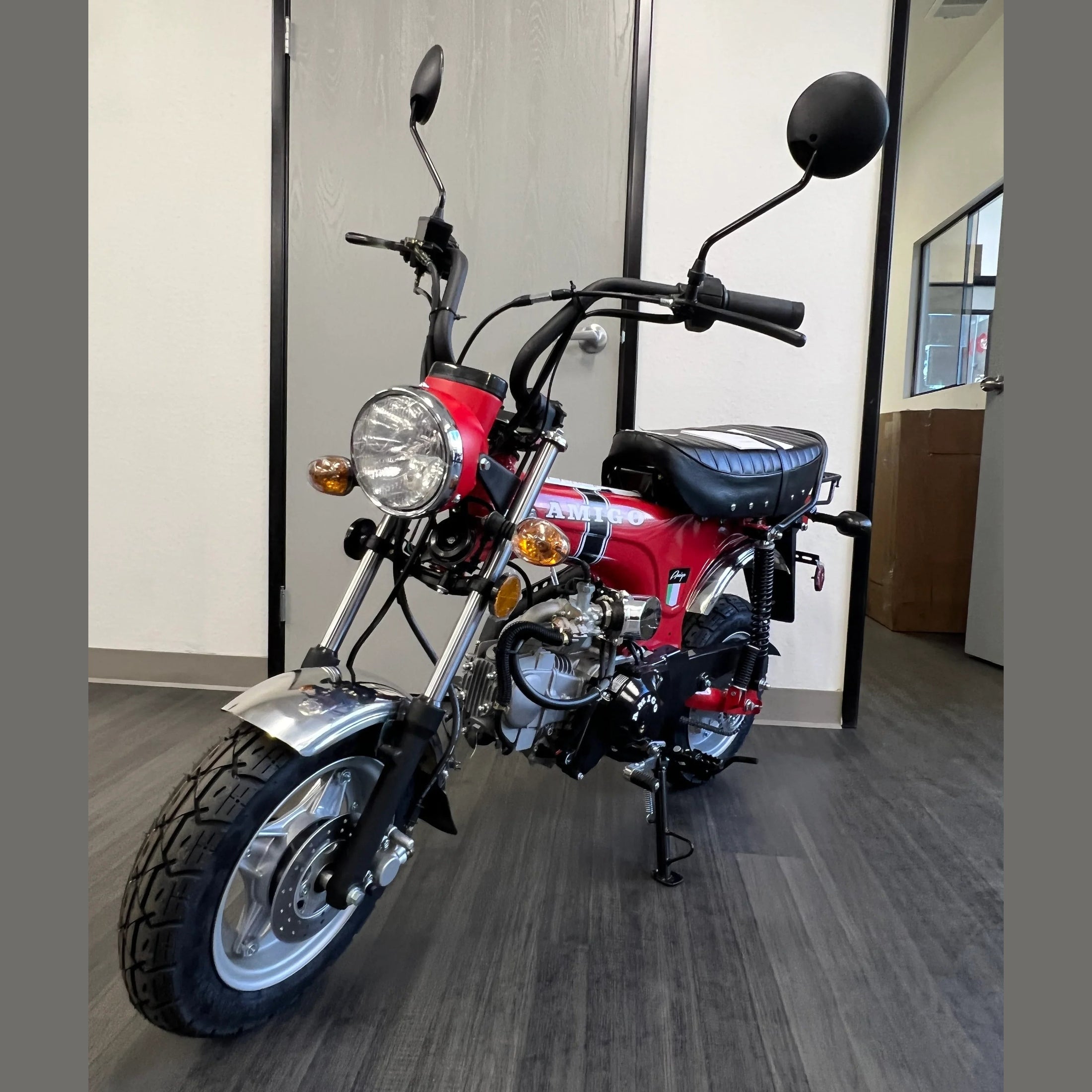 Red Amigo mini bike close-up showing exposed engine, chrome forks, and leather-style seat