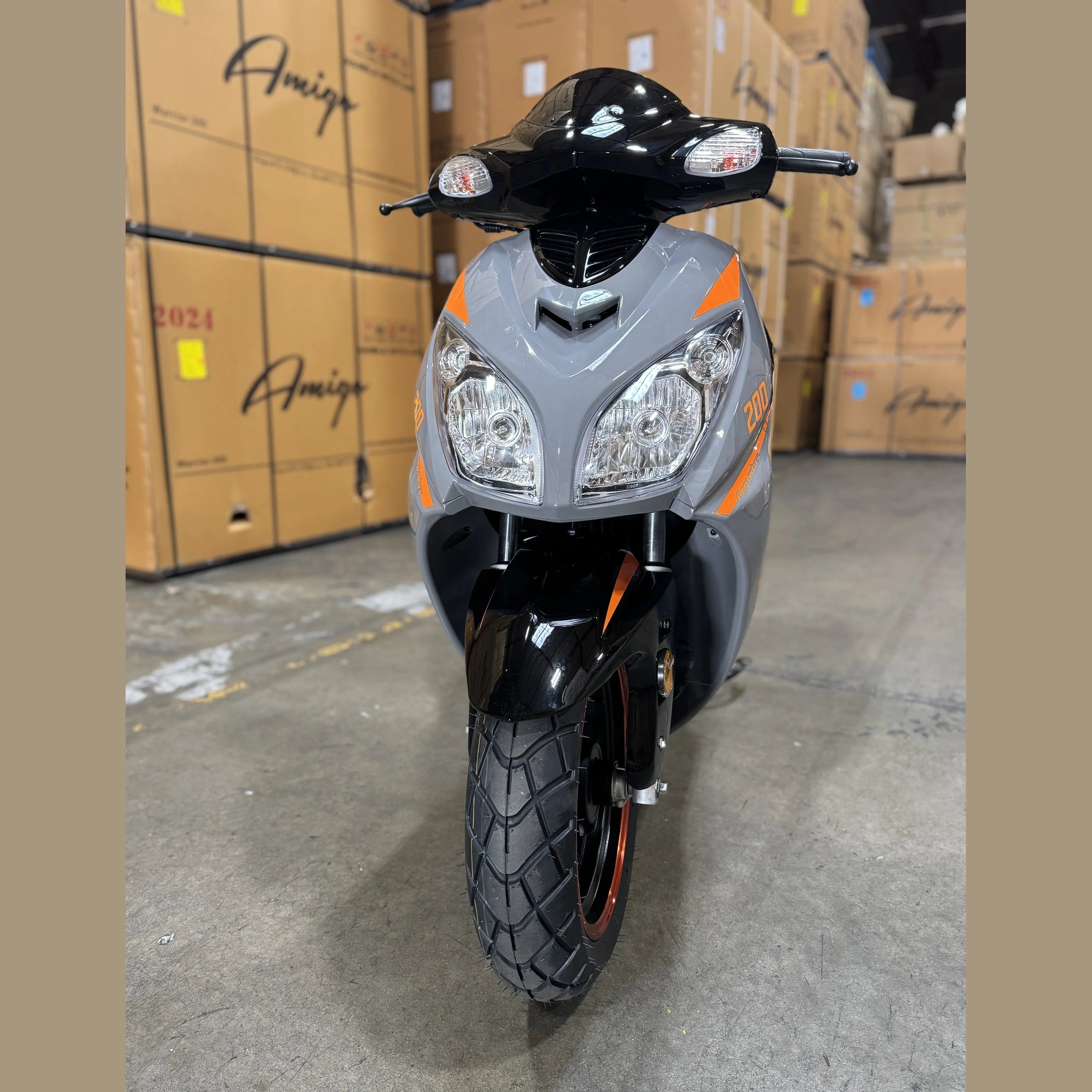 Gray and black scooter with dual headlights in a warehouse, surrounded by shipping boxes.