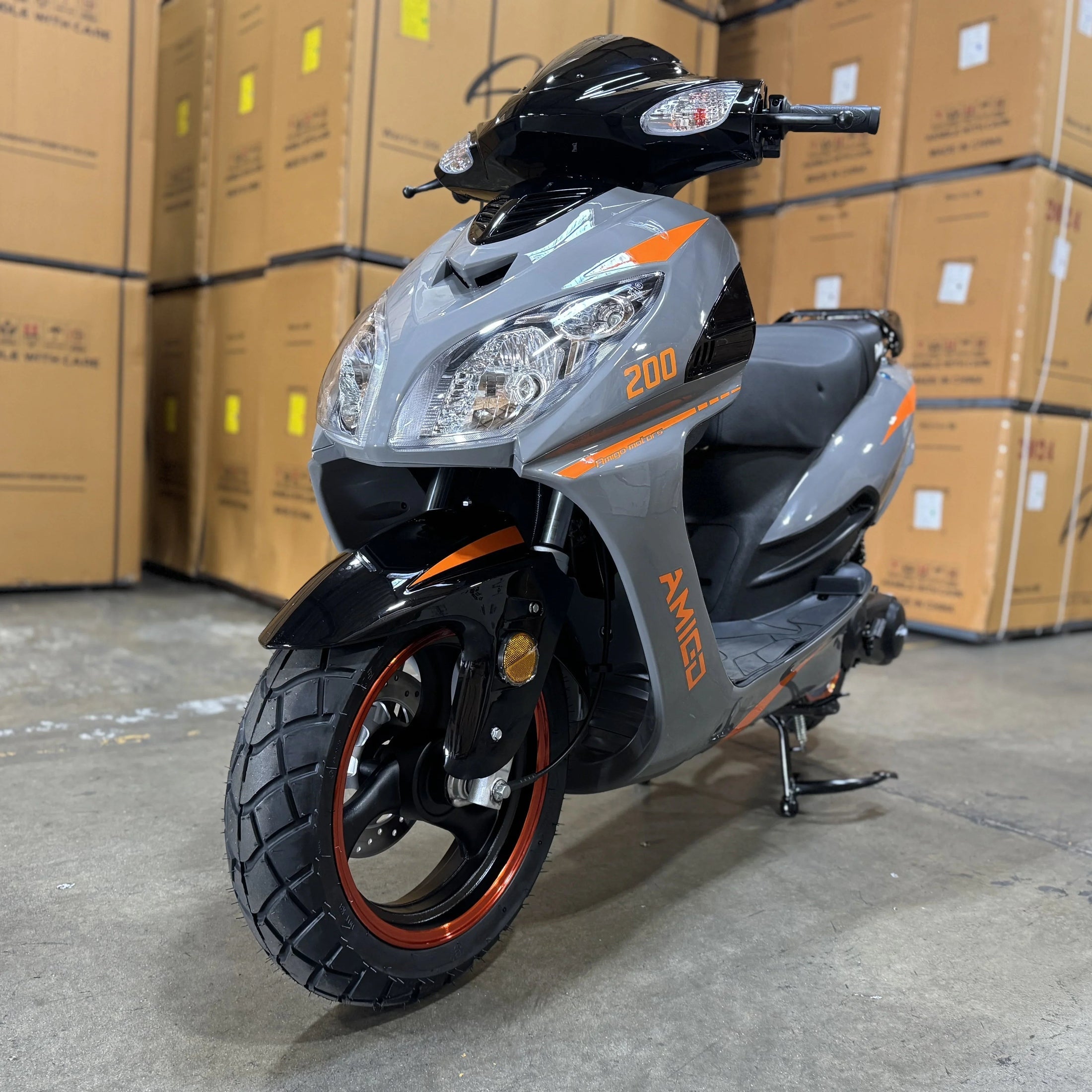 Gray Amigo scooter with orange accents parked in warehouse, stacked boxes in background