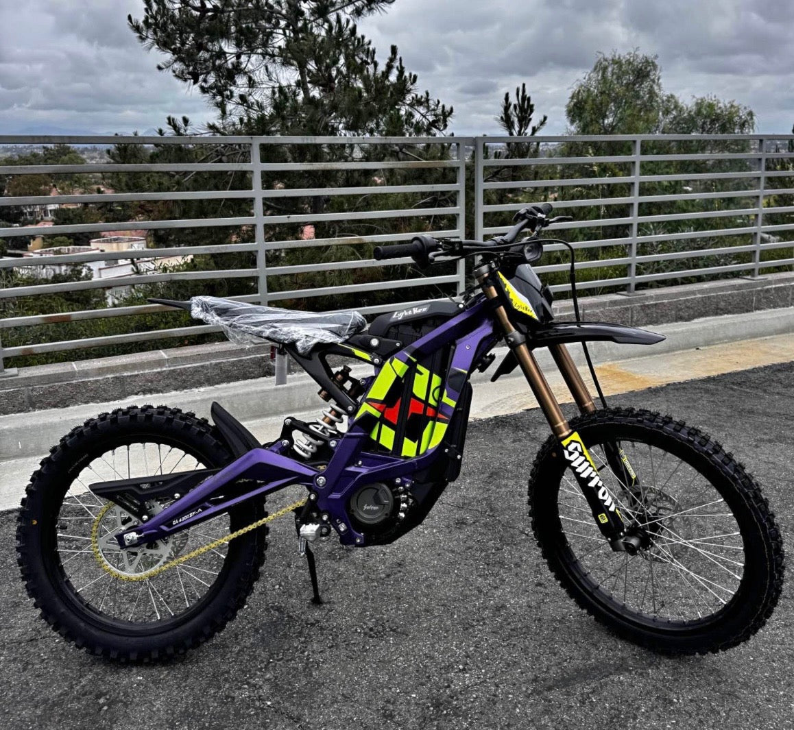 Purple surron light bee LBX electric bike with yellow and black accents on a road with a metal fence and trees in the background.