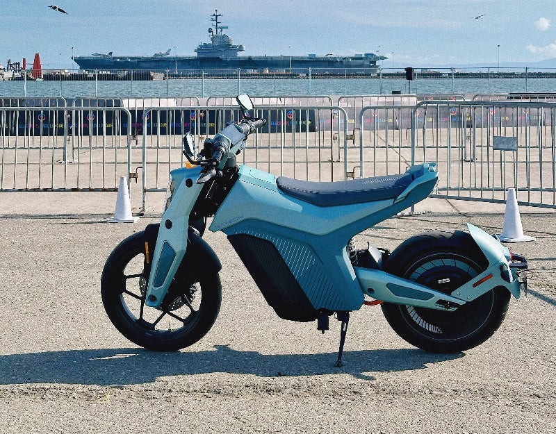 Light blue Naxeon IAM electric motorcycle parked on pavement by waterfront with aircraft carrier and clear sky in background