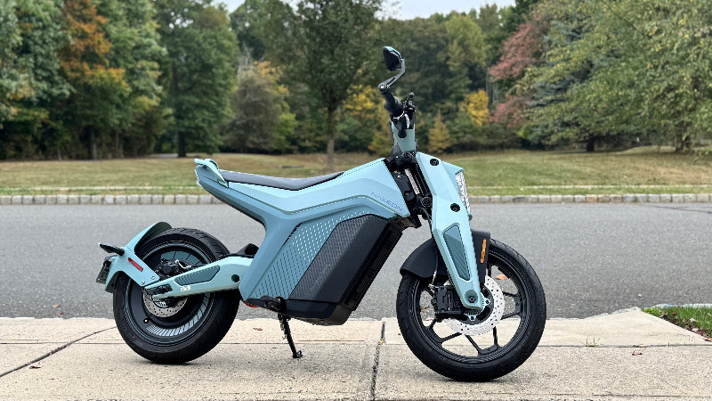 Light blue Naxeon electric scooter parked on sidewalk with a park and trees in the background