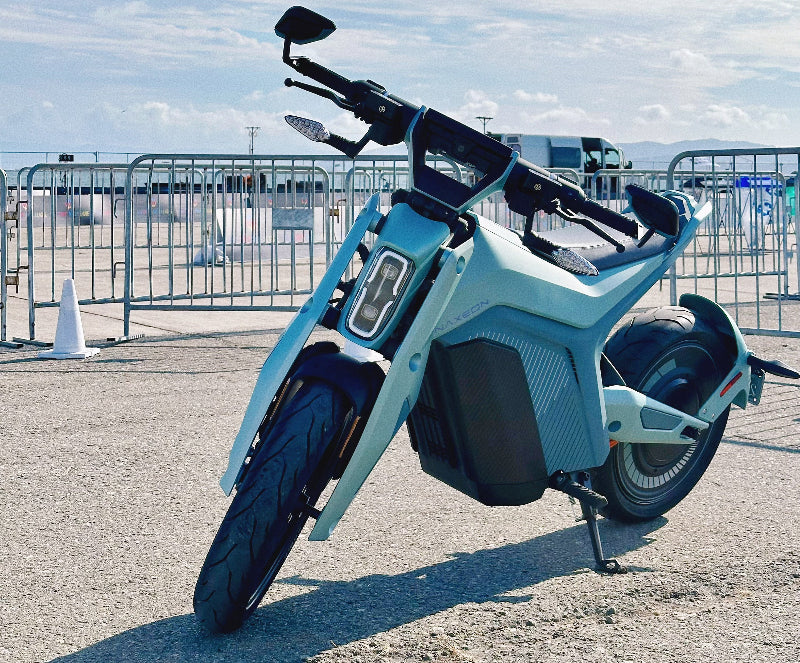 Light blue Naxeon electric motorcycle parked on asphalt with clear sky and metal barriers in background