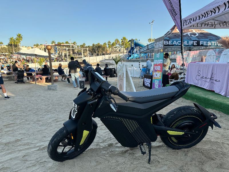 Matte black electric Naxeon IAM motorcycle on sandy beach with festival tents and crowd in background