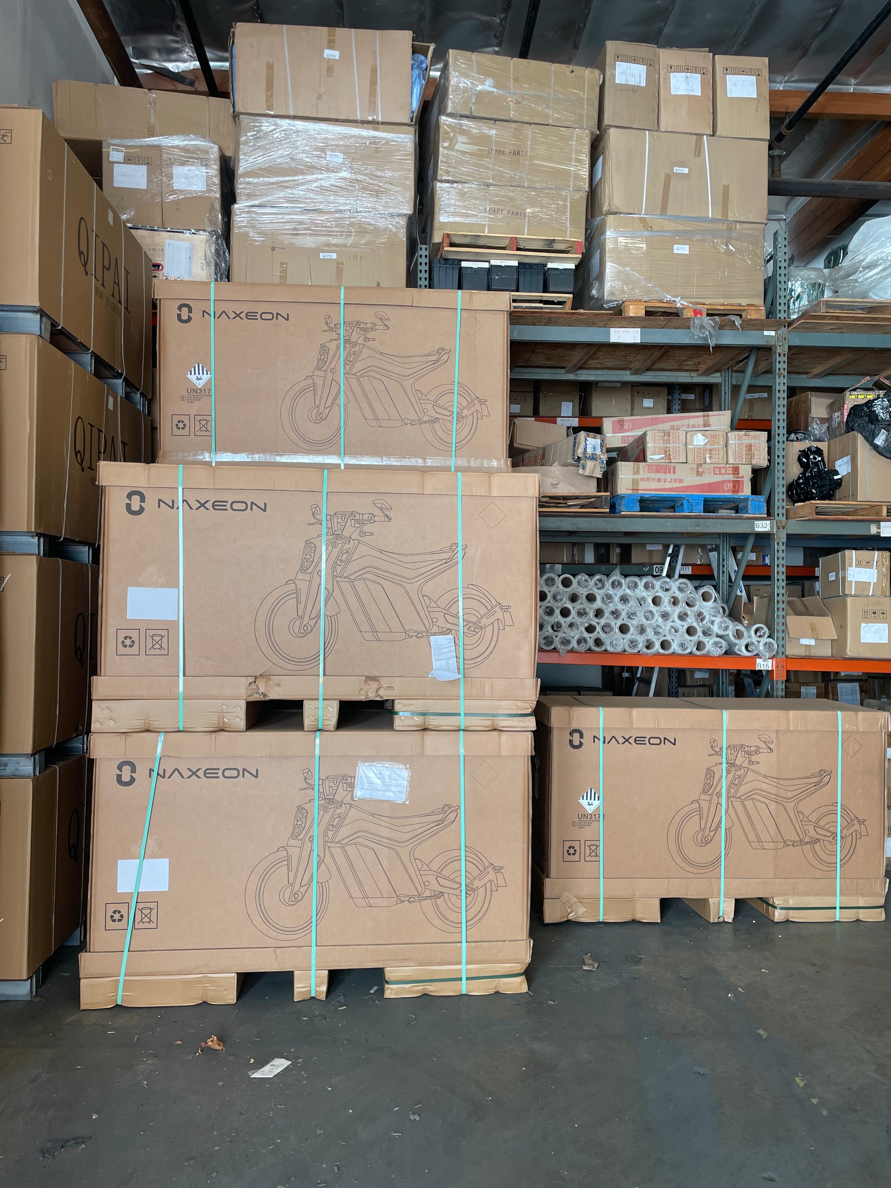 Warehouse stacked with large cardboard boxes labeled Naxeon, containing electric motorcycles, surrounded by additional boxed parts and inventory