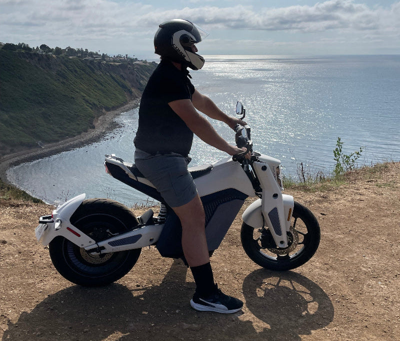 Man wearing black helmet and shorts sitting on White Naxeon IAM Electric Motorcycle overlooking coastal cliff and ocean