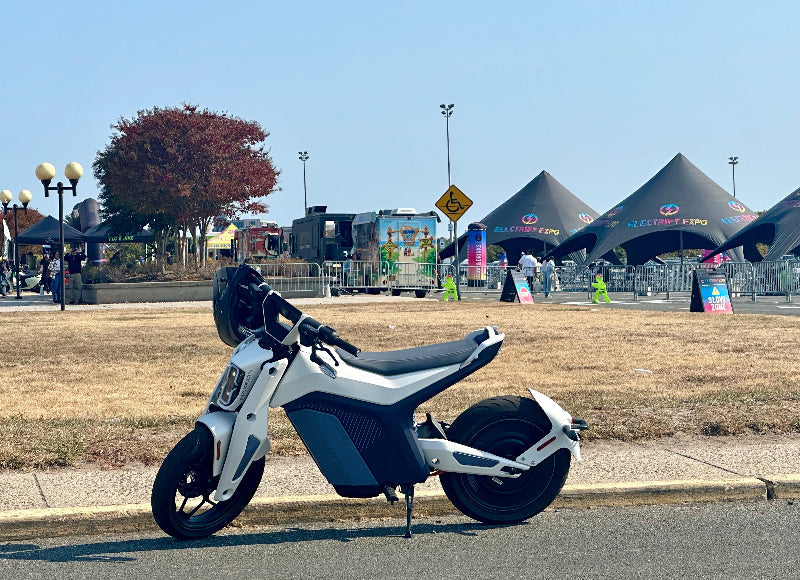 White and black Naxeon electric motorcycle parked on street near Electrify Expo tents and food trucks