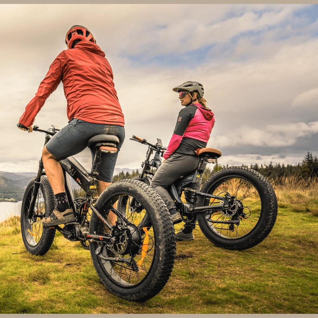 Two riders on black Freesky electric fat tire bikes overlooking lake and forest from grassy hill